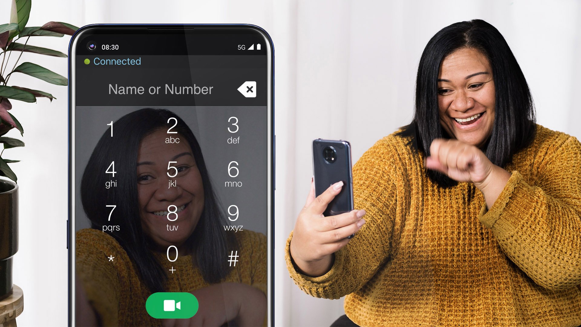  A Polynesian woman with a long bob of black hair and wearing a yellow mustard knitted jumper, holding a phone while video chatting. She is standing in front of an off-white curtain and an indoor plant. On the left side of the image is a demo display of the Convo Australia app.