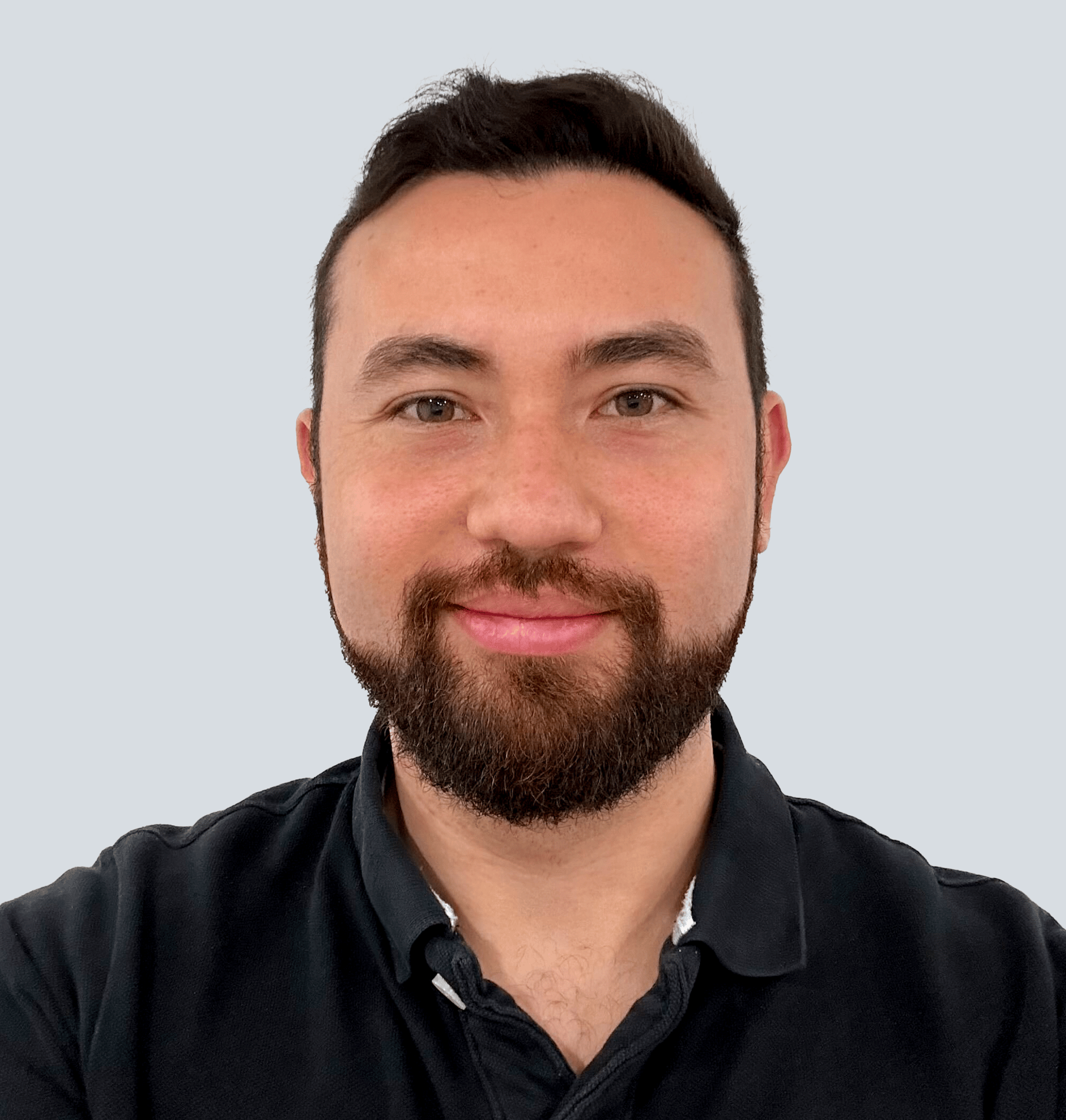 Olwyne, a Caucasian man with dark brown hair and a short cropped dark brown beard and he is wearing a black polo shirt. He is standing in front of a light grey wall.