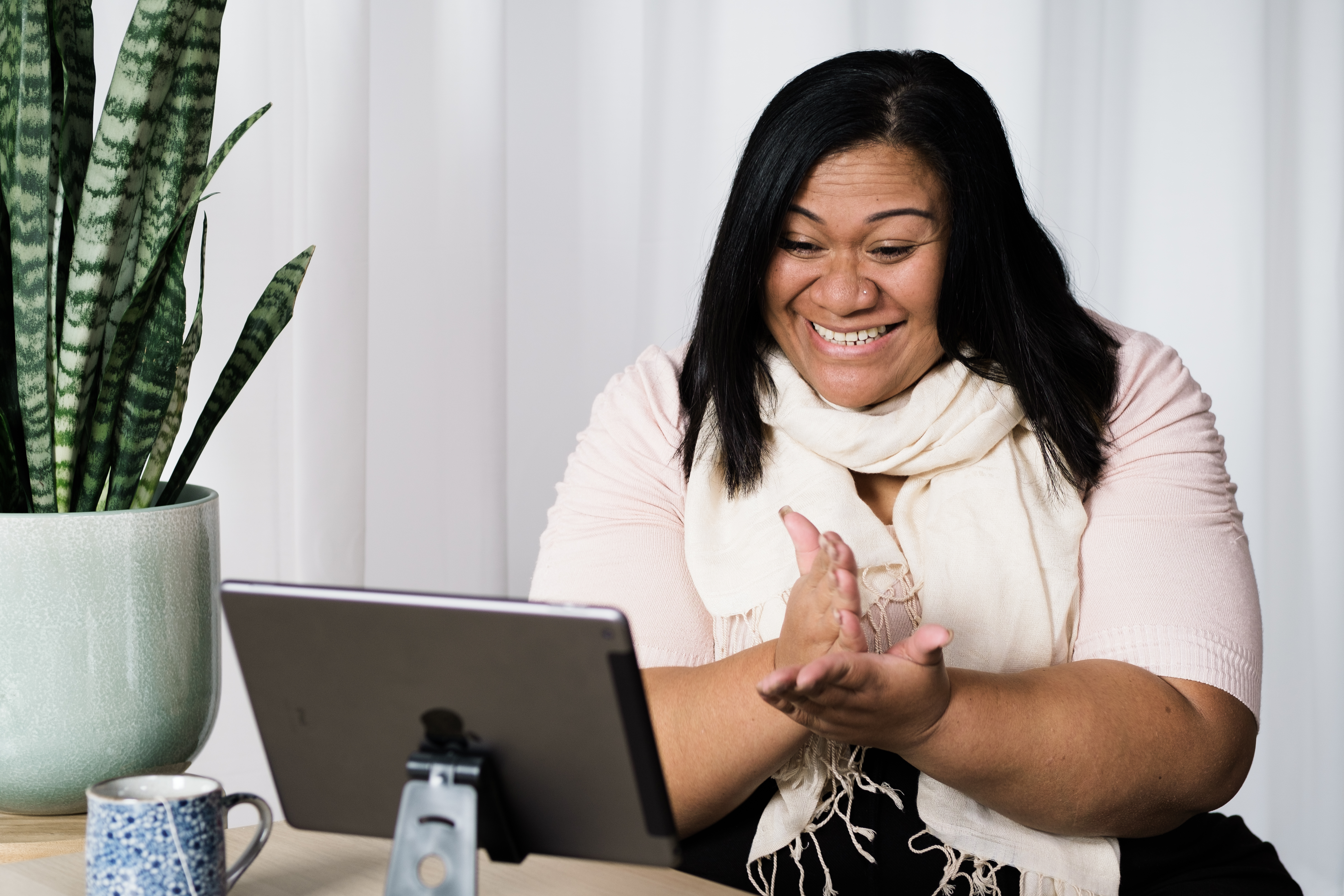 A Polynesian woman with a stylish long bob of black hair is wearing a light pink top and a white scarf. She is using sign language to communicate with someone through a video chat on her tablet. The woman is standing in front of an off-white curtain and an indoor plant, creating a pleasant and inviting background for the video call.