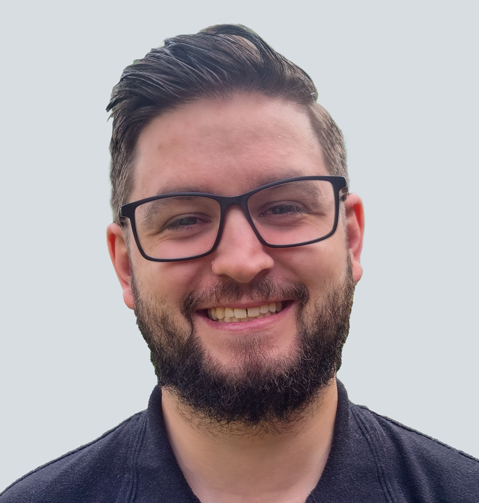 Joshua, a Caucasian man with dark brown hair and a short cropped dark brown beard, wearing black framed glasses and a black polo shirt. He is standing in front of a light grey wall.