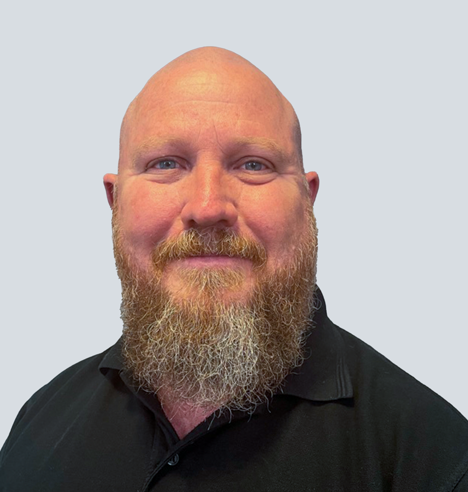 Kyle, a Caucasian man with a shaved head, medium-length ginger and grey beard, wearing a black polo shirt. He is standing in front of a light grey wall.