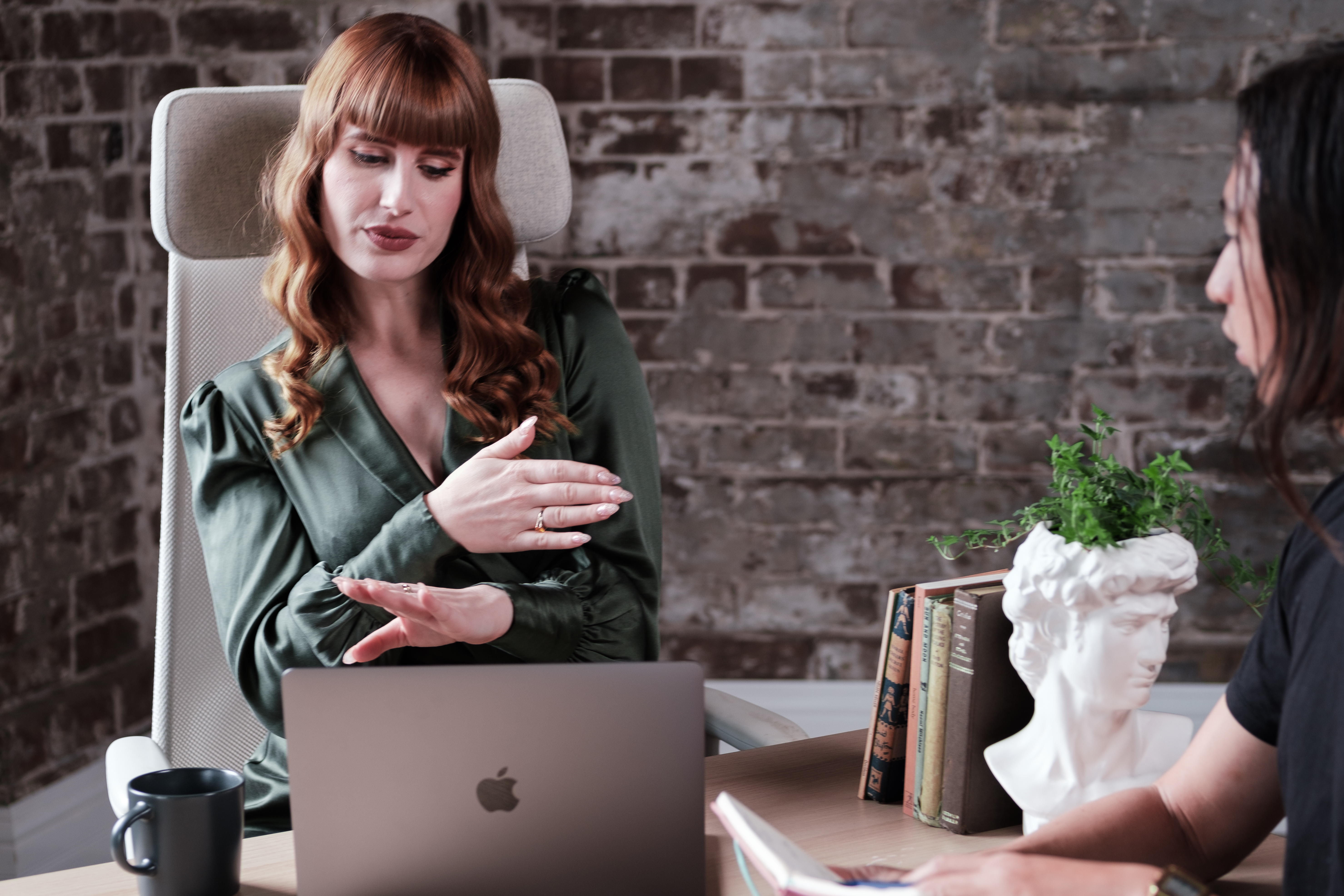 A woman with long red hair is wearing a green silk blouse. She is talking to a colleague who can hear her through a video chat on her laptop. The colleague is shown on the right side of the image. The woman is using sign language to communicate with her colleague during the face-to-face interaction meeting in person.