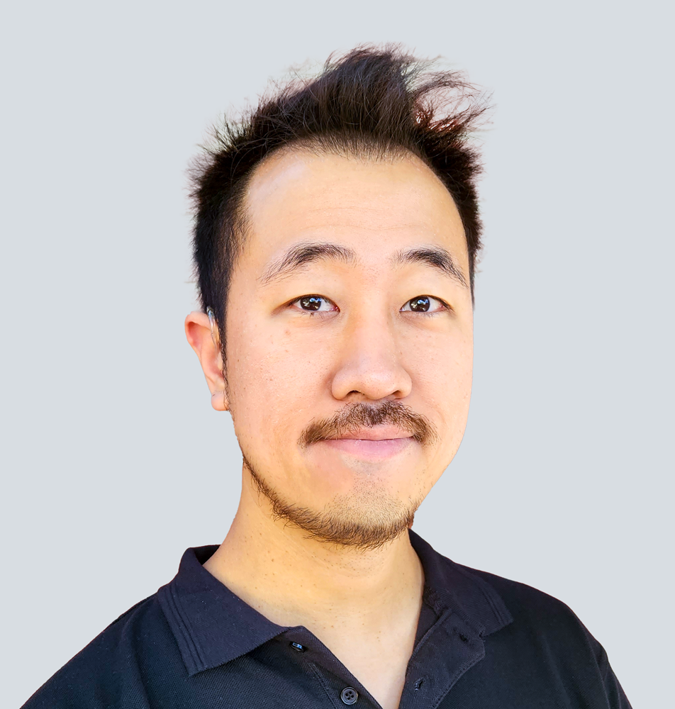 SaengJae, an Asian man with short black hair and facial hair, wearing a black polo shirt. He is standing in front of a light grey wall.