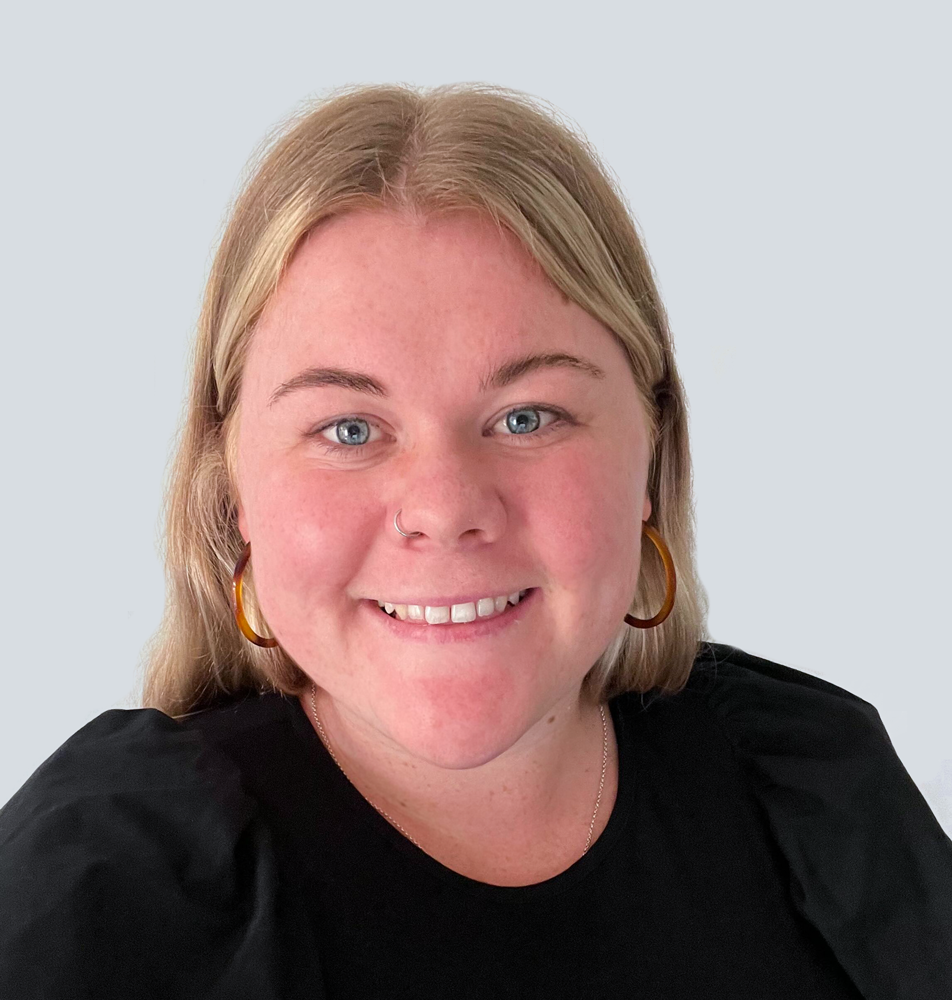 Tiahna, a Caucasian woman with long blonde hair, wearing a black shirt. She is standing in front of a light grey wall.