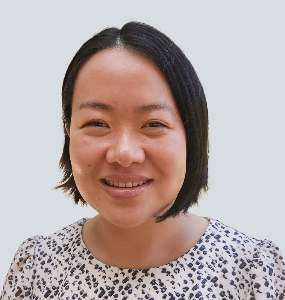 Tina, an Asian woman with long bob black hair, wearing a white top with black dots. She is standing in front of a light grey wall.