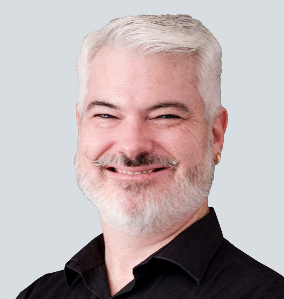 Todd, a Caucasian man with white-grey hair, white-grey beard, and a curly moustache, wearing a black polo shirt. He is standing in front of a light grey wall.